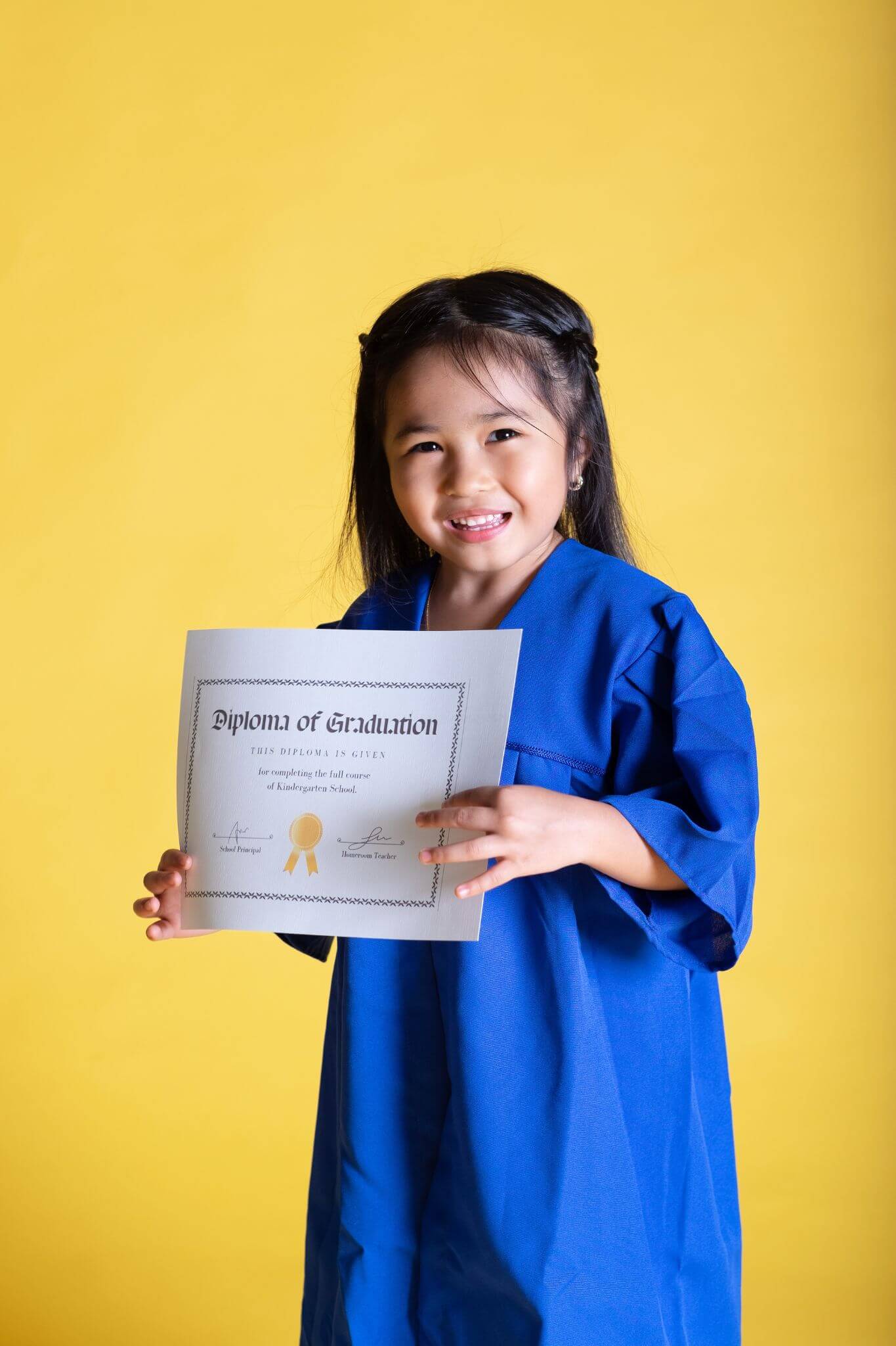 A kindergarten-age girl smiles in her graduation gown holding her diploma from kindergarten graduation